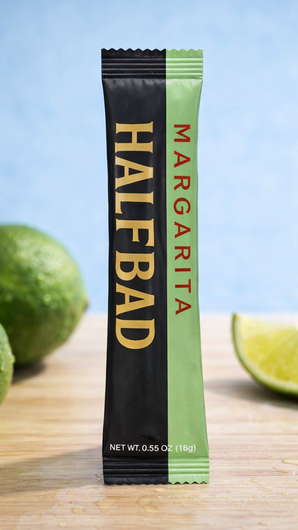 Half Bad Margarita packets with limes and a lime wedge on a light blue background